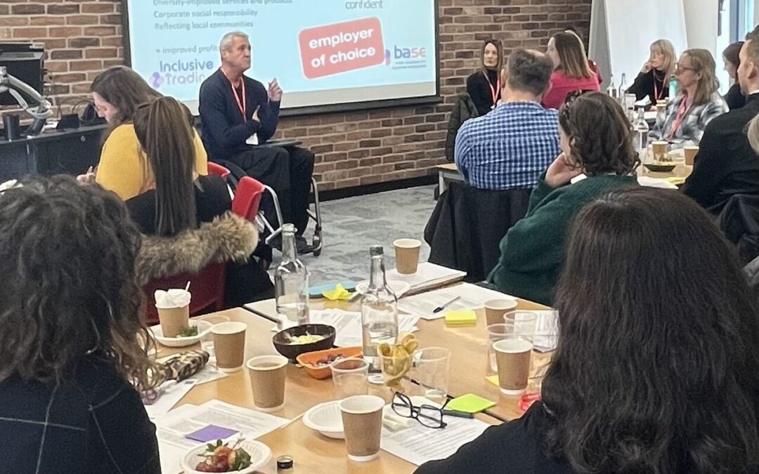 Photo is taken from the back of a room where a workshop is being delivered. Groups of 8 people sit around tables whilst a man in a wheelchair is talking to them.